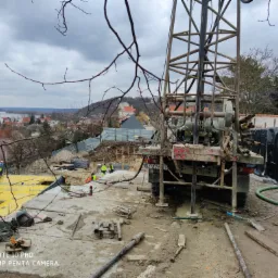 Wiertnica na placu budowy z widokiem na okoliczne budynki, prace w toku, widoczni pracownicy w odblaskowych kamizelkach i kaskach, w tle panorama miasta pod pochmurnym niebem.