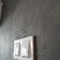 Szara ściana o wyraźnej, chropowatej fakturze z zamontowanym białym podwójnym włącznikiem światła marki Simon.