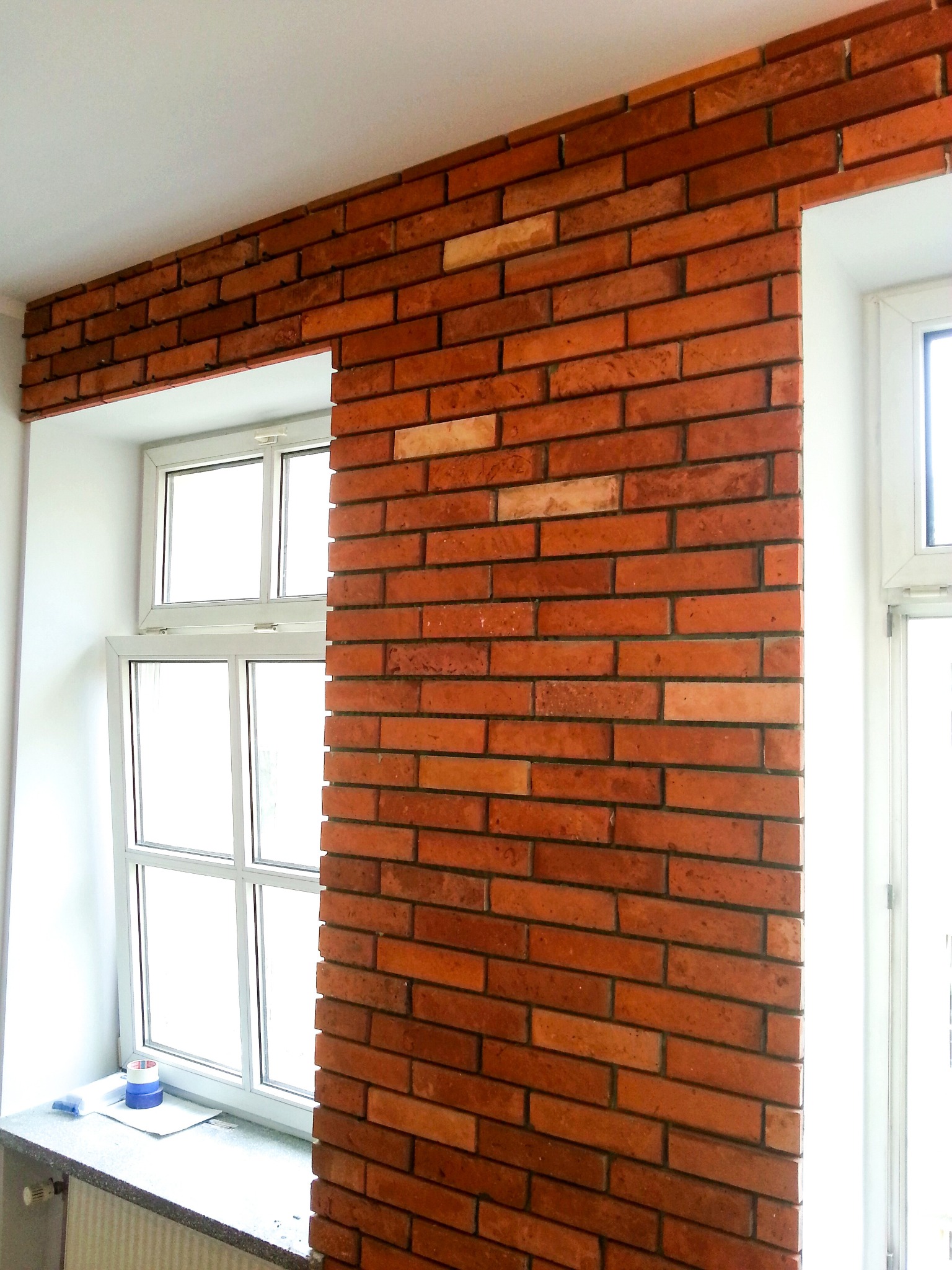 Ściana wykończona czerwoną cegłą klinkierową, obramowująca okno z białymi ramami. Na parapecie leży niebieska taśma malarska.