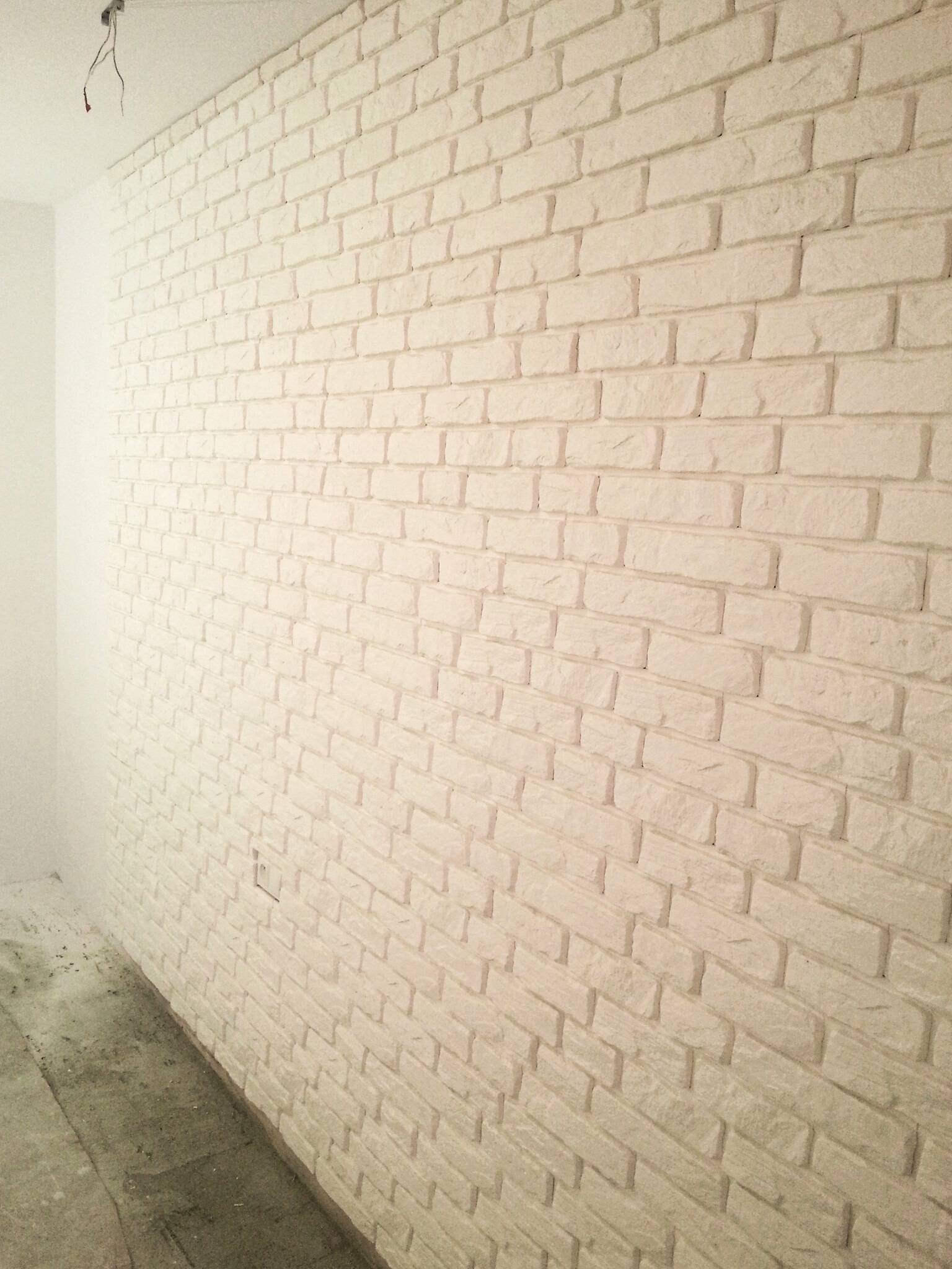 Ściana wykończona białymi płytkami imitującymi cegłę, widoczne przewody elektryczne wystające z sufitu, surowa podłoga w trakcie prac wykończeniowych.