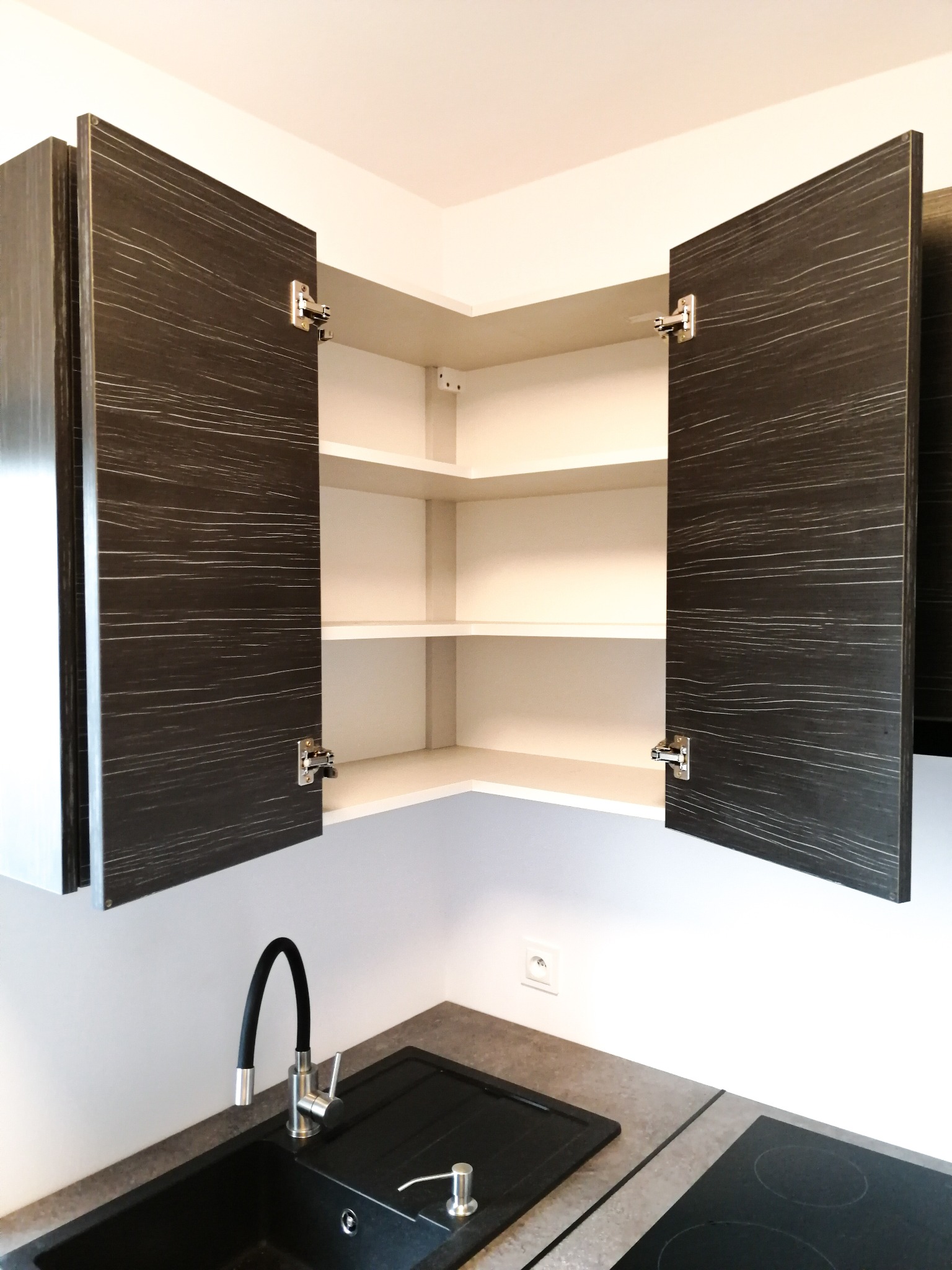 Kątowa szafka kuchenna z otwartymi drzwiczkami o fakturze ciemnego drewna, widok na puste, białe półki, poniżej czarny zlew z baterią i dozownikiem na płyn.
