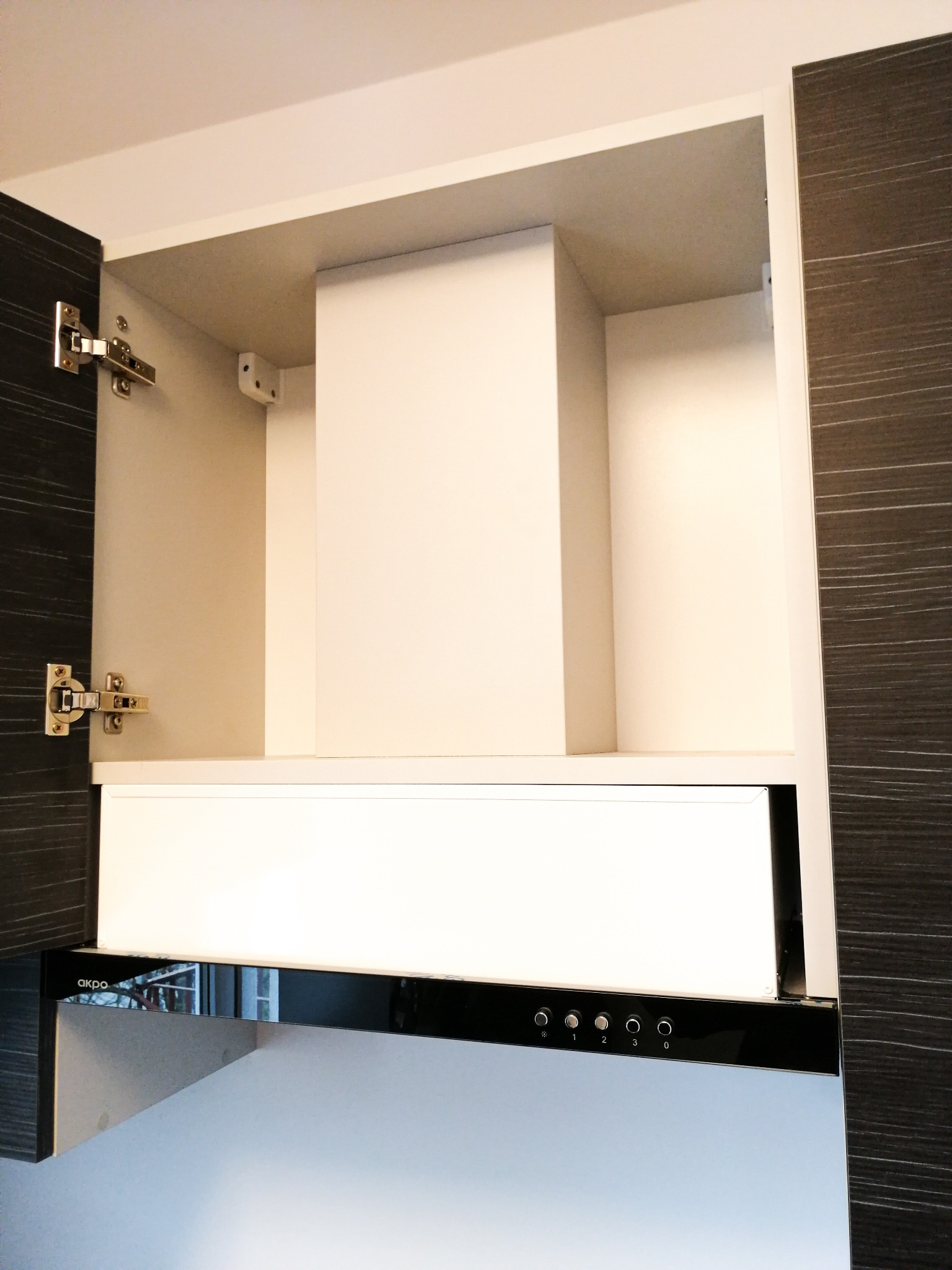 Wnętrze szafki kuchennej z zamontowanym okapem marki Akpo, widoczne zawiasy i panel sterowania okapu.