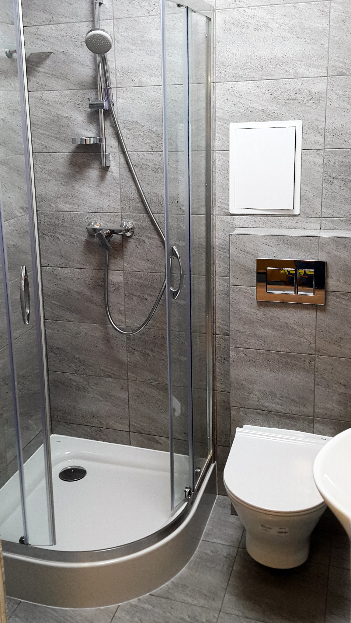 Wykończona łazienka z kabiną prysznicową z przesuwnymi drzwiami, toaletą podwieszaną z chromowanym przyciskiem spłukującym i szarymi płytkami imitującymi kamień na ścianach i podłodze.