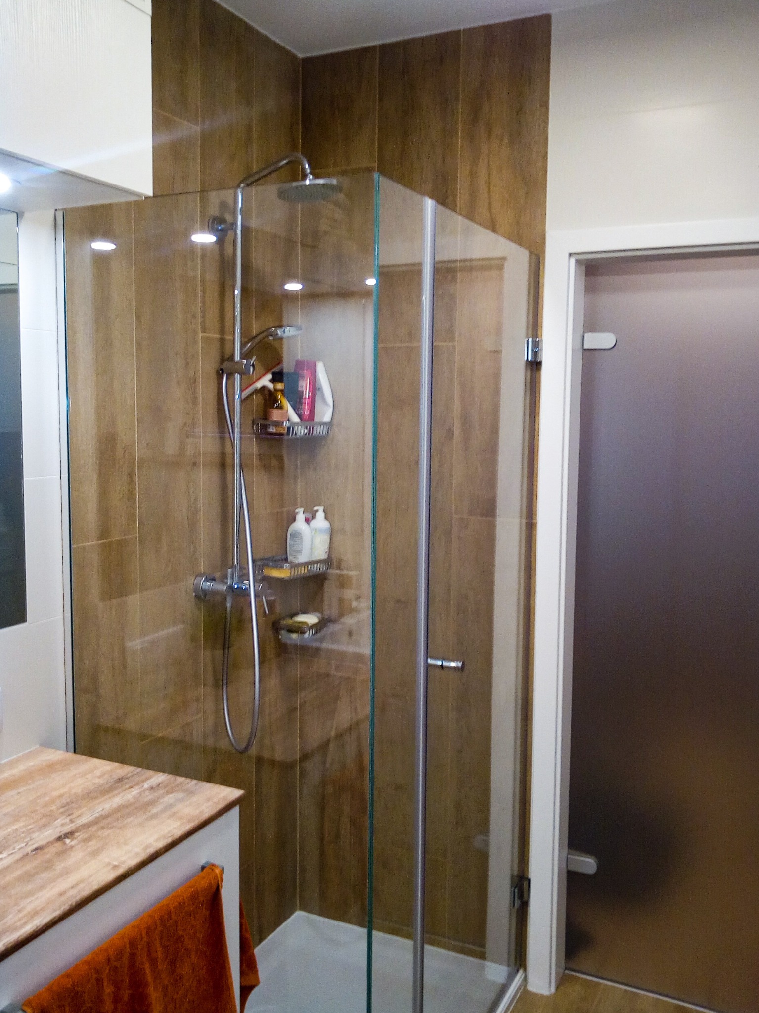 Nowoczesna kabina prysznicowa z brązowymi płytkami imitującymi drewno, wyposażona w półki z kosmetykami i chromowaną armaturę, widoczna umywalka z blatem o wzorze marmuru i pomarańczowy ręcznik.