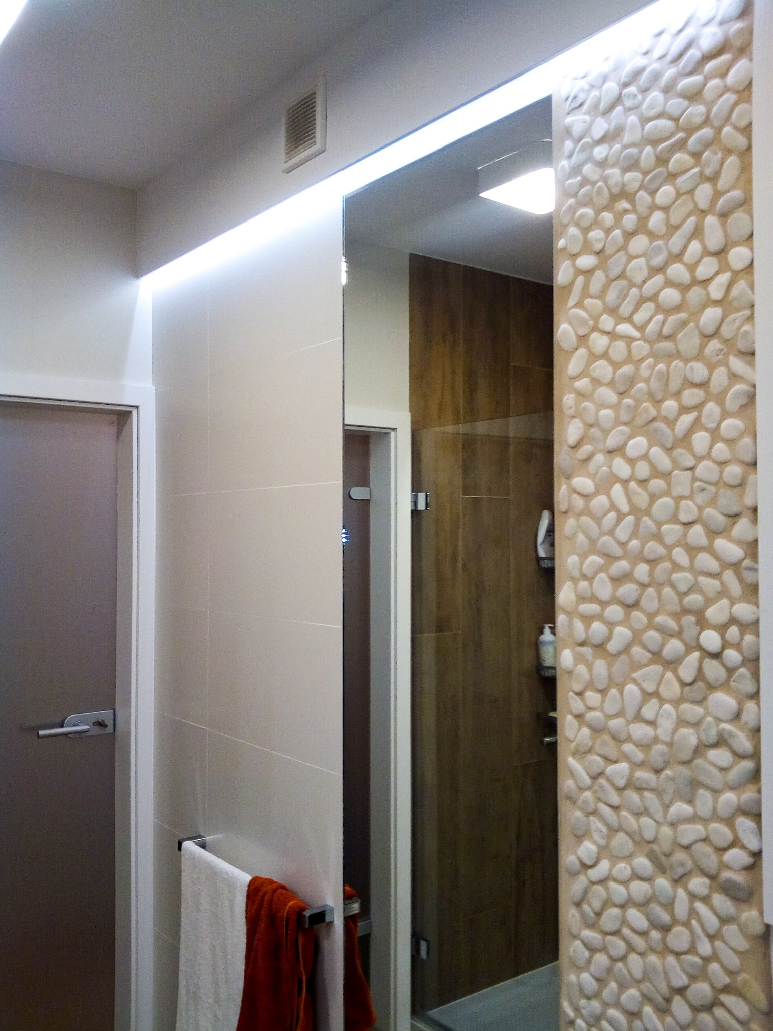 Nowoczesna łazienka z ścianą wyłożoną dekoracyjnymi kamieniami rzecznymi, podświetleniem LED, szklaną kabiną prysznicową i ręcznikami w odcieniach bieli i terakoty.