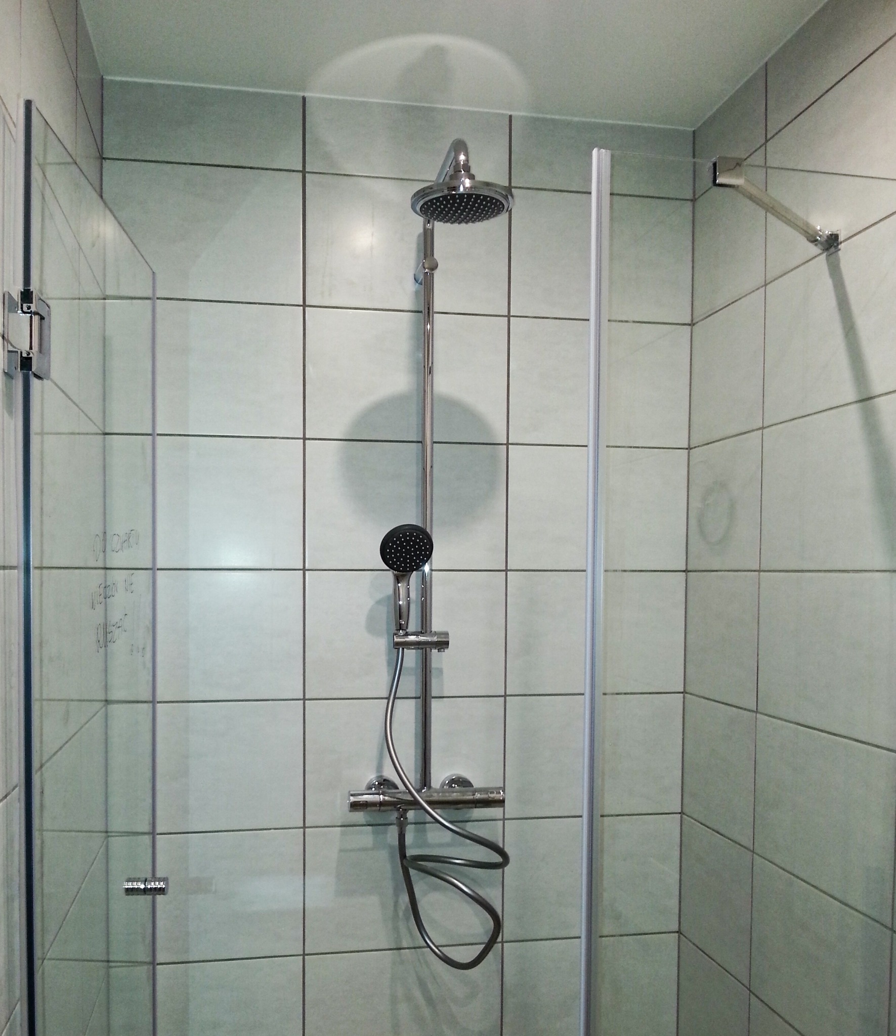 Nowoczesna kabina prysznicowa z chromowaną armaturą, szklaną ścianką i szarymi płytkami; na szklanej ściance widoczne zacieki i napisy.