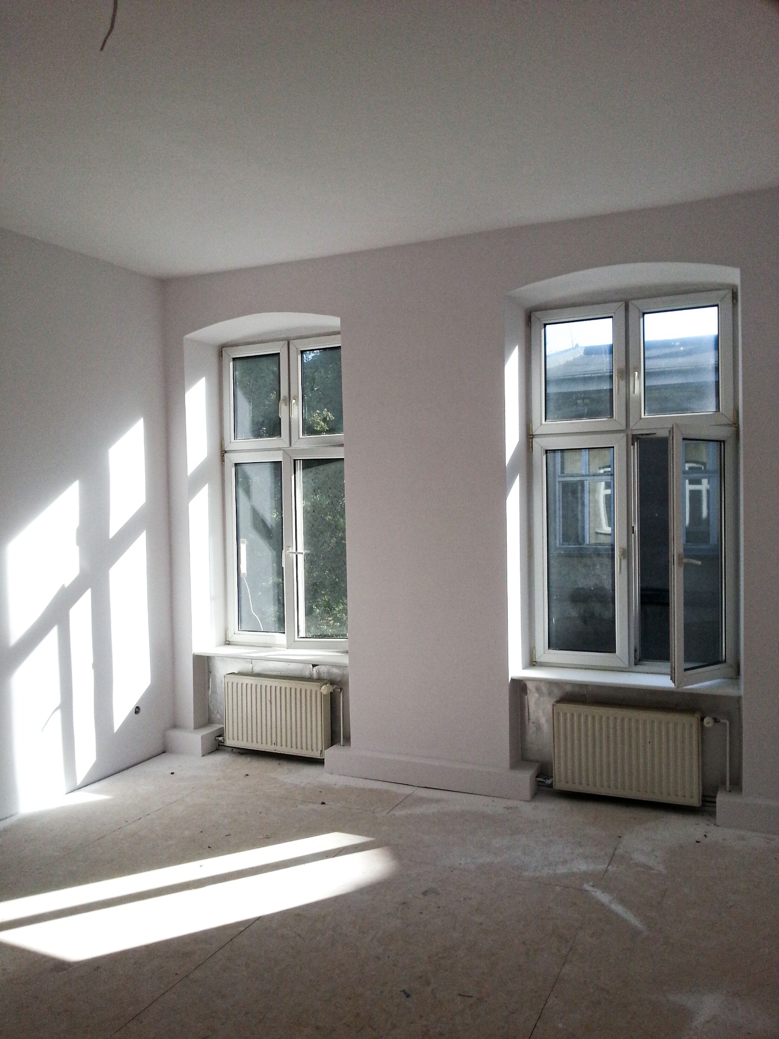 Jasne, puste pomieszczenie z dwoma oknami z białymi ramami, świeżo pomalowane ściany i grzejniki pod oknami, widoczne ślady po pracach wykończeniowych na podłodze, promienie słońca wpadają przez okno.