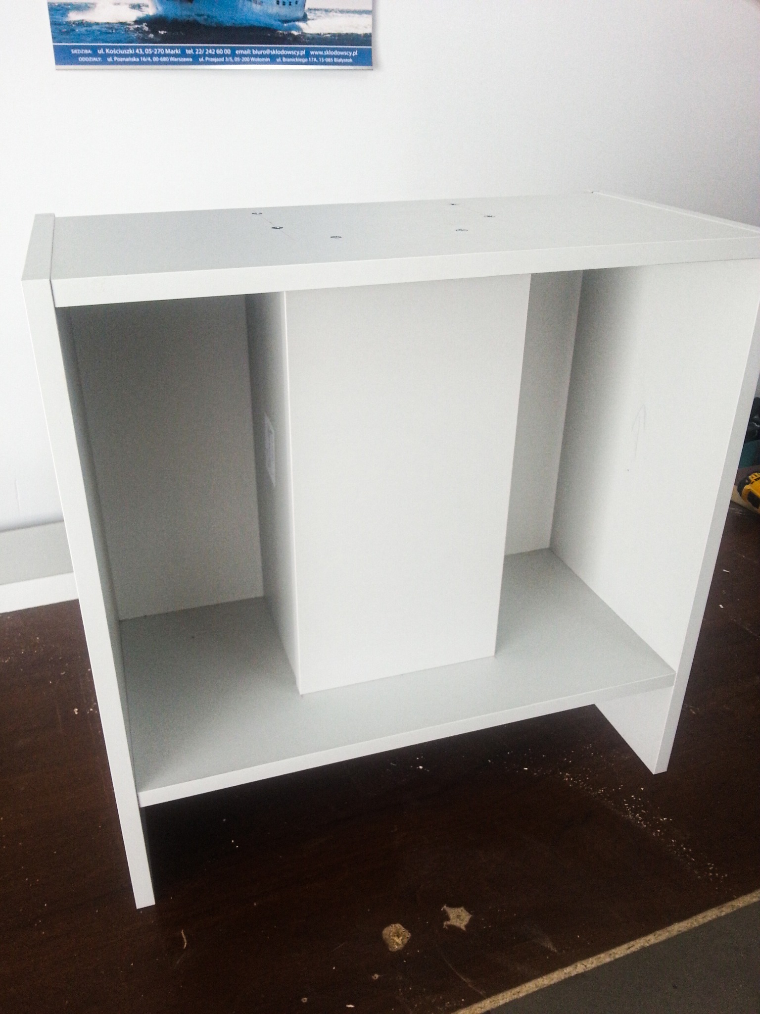 Biała, częściowo zmontowana szafka łazienkowa z dwiema półkami, widoczna na tle ściany i fragmentu podłogi, z plakatem firmy na ścianie w tle.