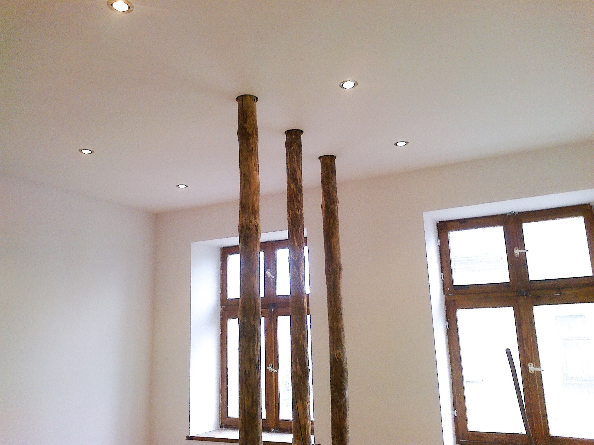 Wnętrze pokoju z trzema surowymi, drewnianymi kolumnami sięgającymi sufitu, w tle okna z drewnianą ramą i wpuszczone oświetlenie sufitowe.