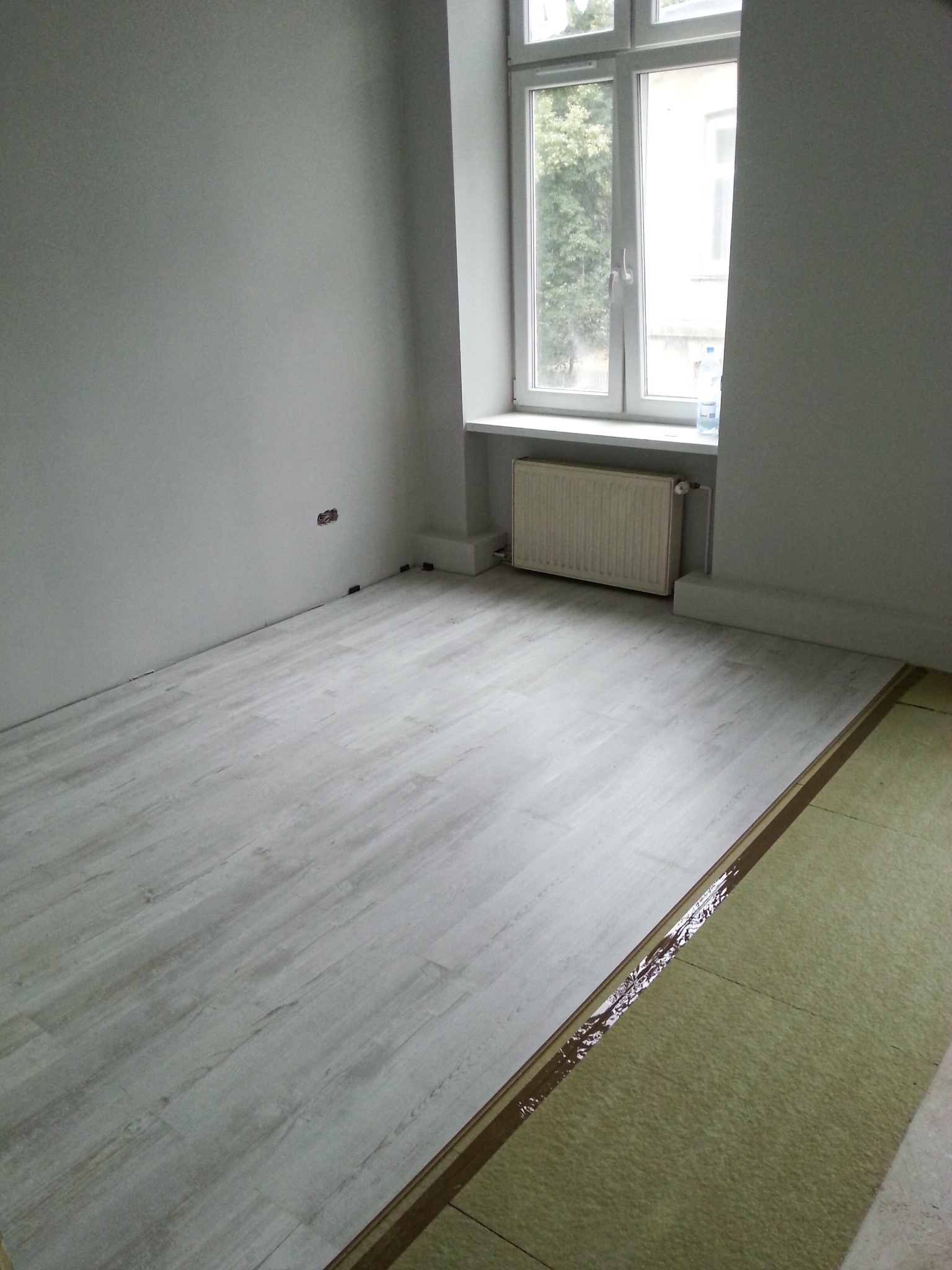 Układanie paneli podłogowych w jasnym odcieniu drewna w pokoju z szarymi ścianami, widoczna warstwa izolacyjna i listwy dystansowe przy ścianie.