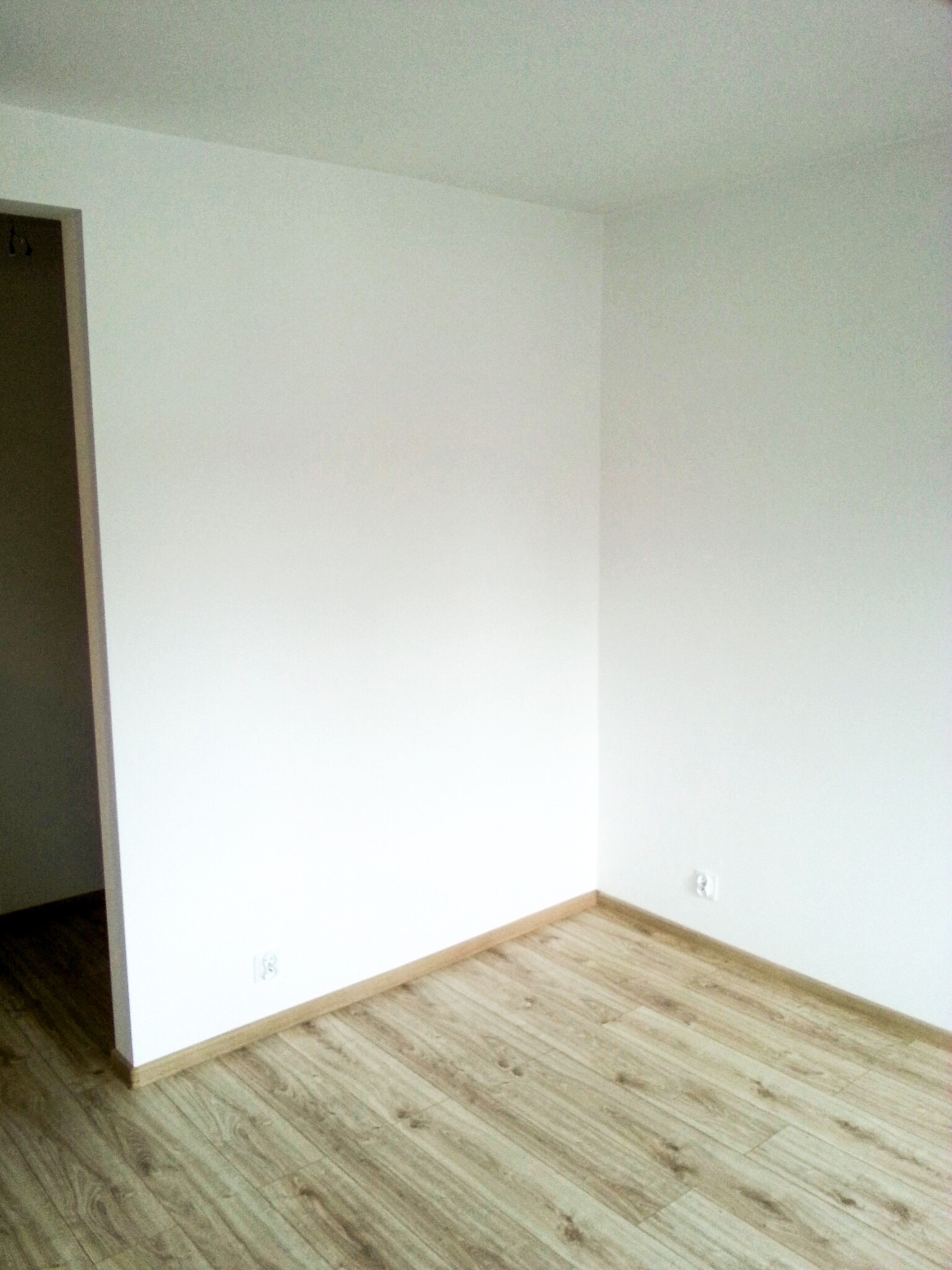 Pusty, świeżo odnowiony pokój z panelami podłogowymi imitującymi drewno, białymi ścianami, widocznymi gniazdkami elektrycznymi i fragmentem futryny drzwiowej.