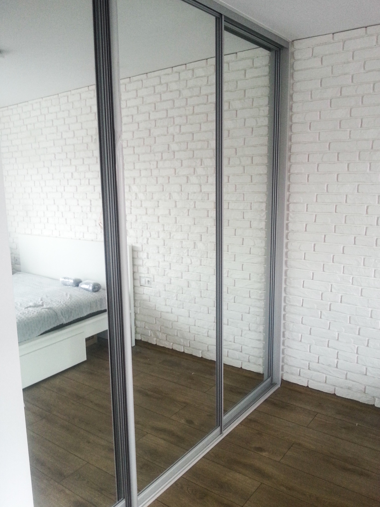 Nowoczesna szafa przesuwna z lustrzanymi drzwiami, odbijająca fragment sypialni z białym łóżkiem, na tle ściany z białej cegły i drewnianej podłogi.