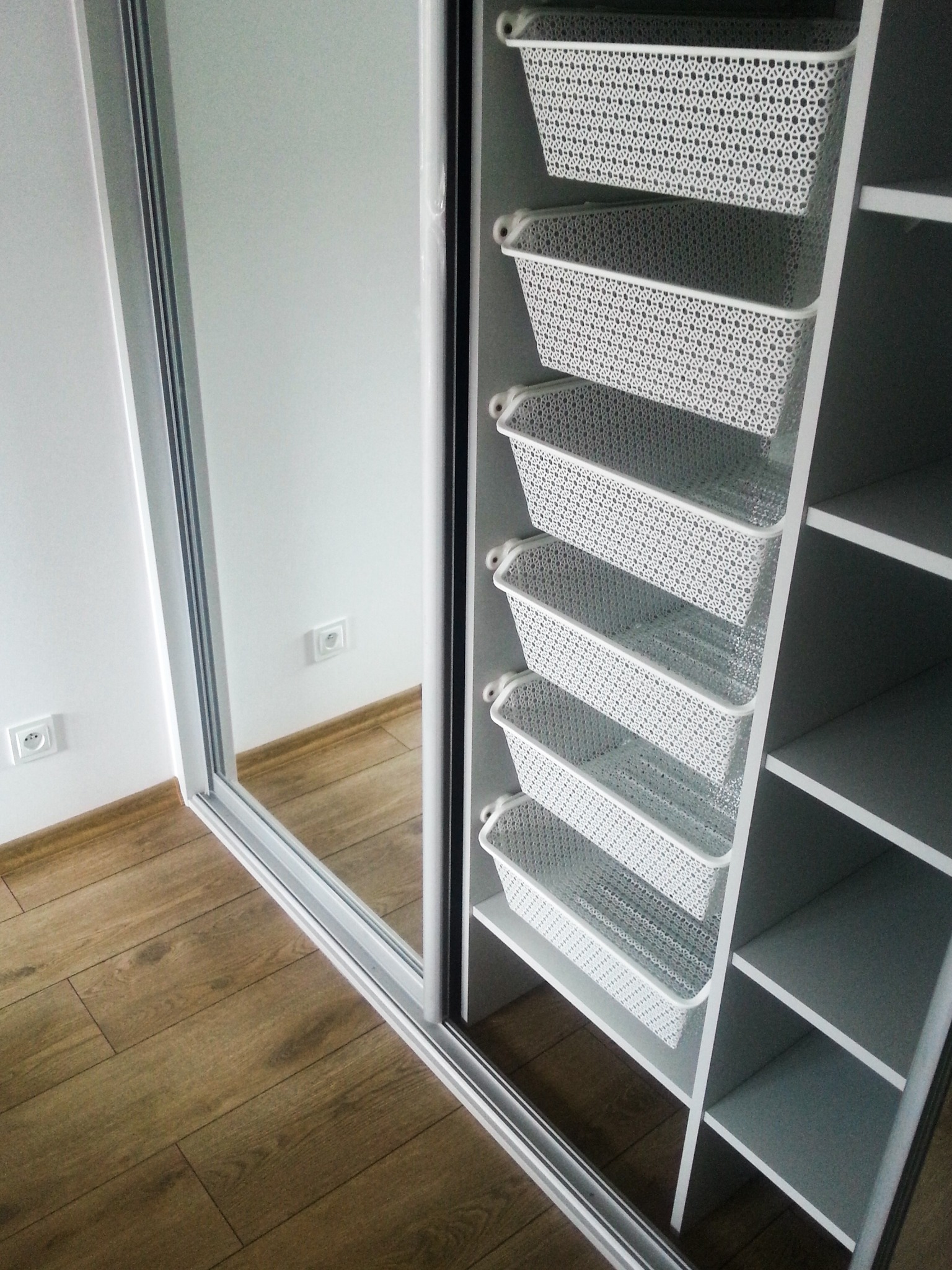 Wnętrze szafy wnękowej z przesuwnymi drzwiami lustrzanymi, zorganizowane za pomocą rzędu białych, ażurowych koszyków na metalowych prowadnicach, widoczne gniazdka elektryczne na białej ścianie.