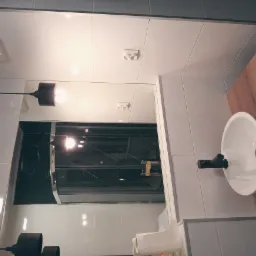 Nowoczesna łazienka z białą umywalką nablatową na drewnianym blacie, czarną armaturą i minimalistyczną lampą wiszącą; widoczne białe płytki na ścianach i lustro odbijające kabinę prysznicową.