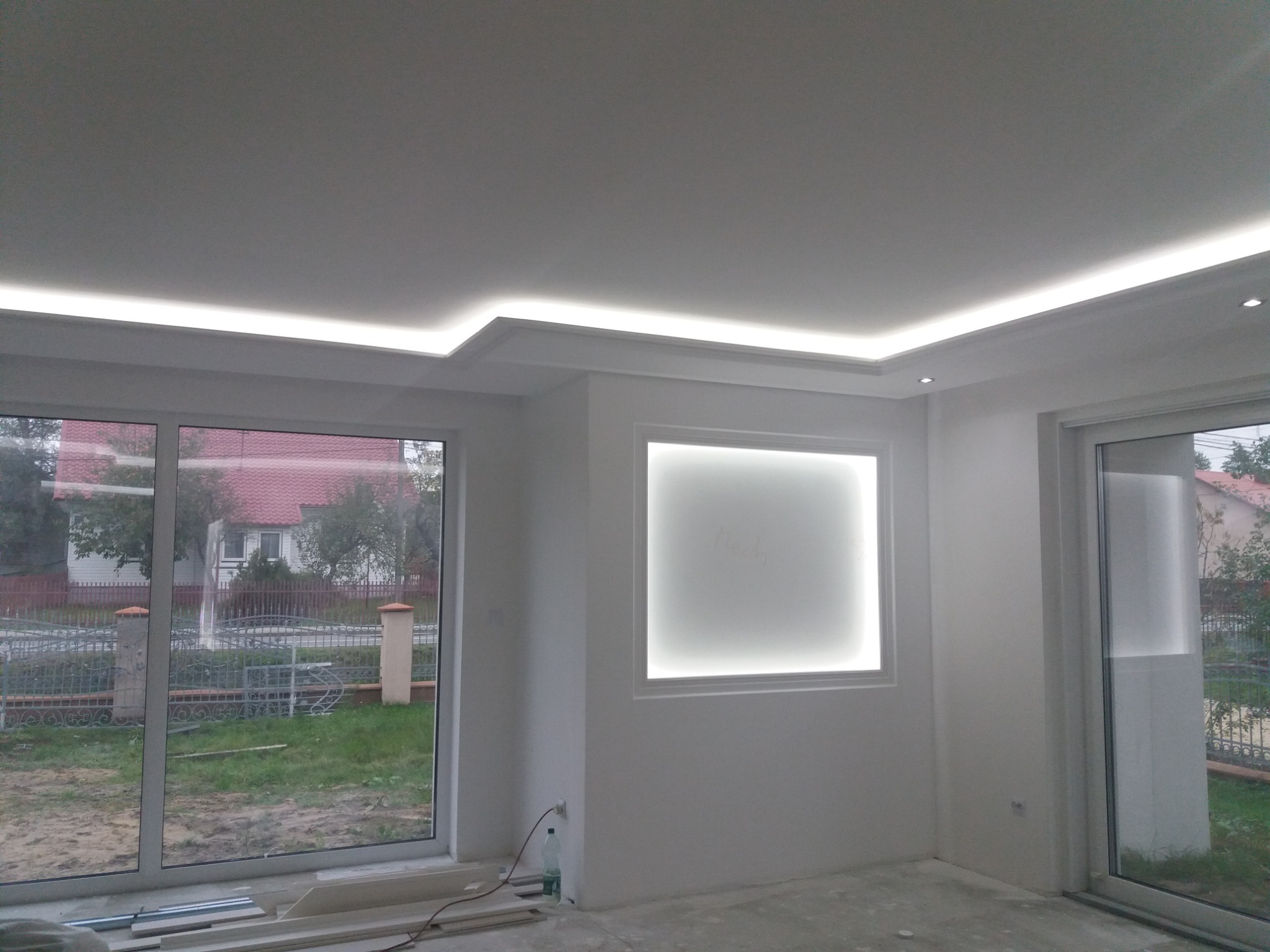 Nowoczesne wnętrze z białymi ścianami, oświetleniem LED w suficie podwieszanym i oświetlonym wnęką na ścianie. Widoczne duże okna z widokiem na zewnątrz.