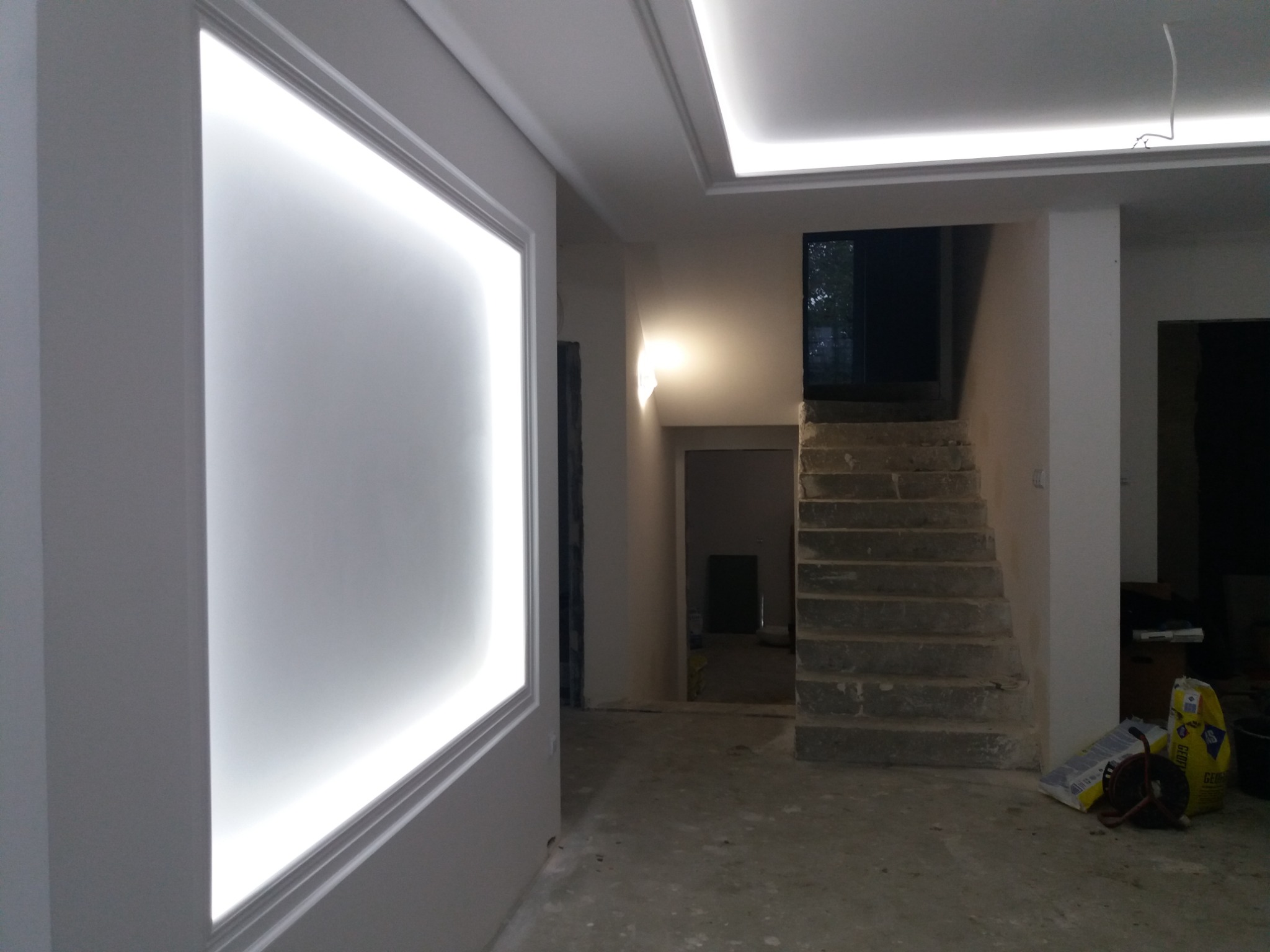 Wnętrze w trakcie remontu: ściana z podświetloną ramą, sufit z oświetleniem LED, surowe schody betonowe i worki z materiałami budowlanymi na podłodze.