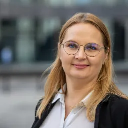 Portret uśmiechniętej blondynki w okularach, ubrana w białą koszulę i czarną marynarkę, na rozmytym tle biurowca.