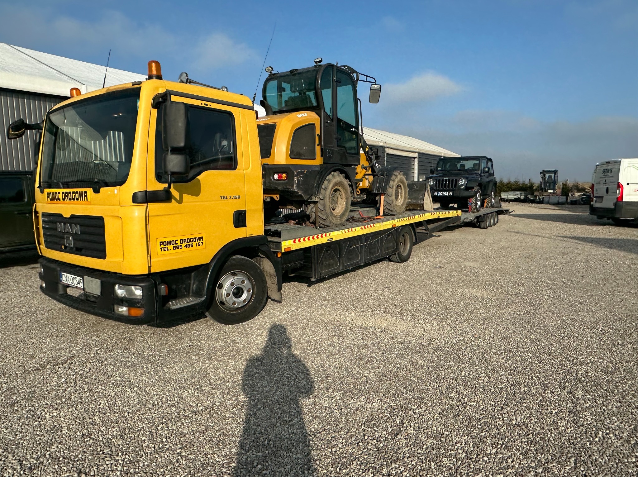 Żółta laweta MAN z koparką WOLF i czarnym Jeepem na platformie, zaparkowana na żwirowym placu w słoneczny dzień. Widoczny cień osoby robiącej zdjęcie.