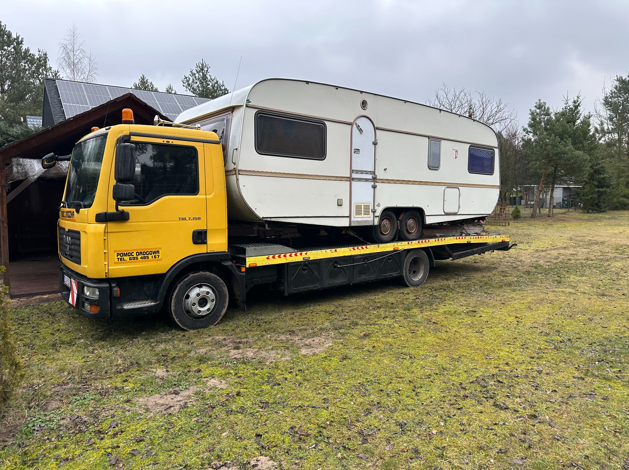 Żółta laweta MAN z przyczepą kempingową na platformie, na tle domu z panelami słonecznymi i drzew. Widoczny napis 'Pomoc Drogowa' na kabinie.