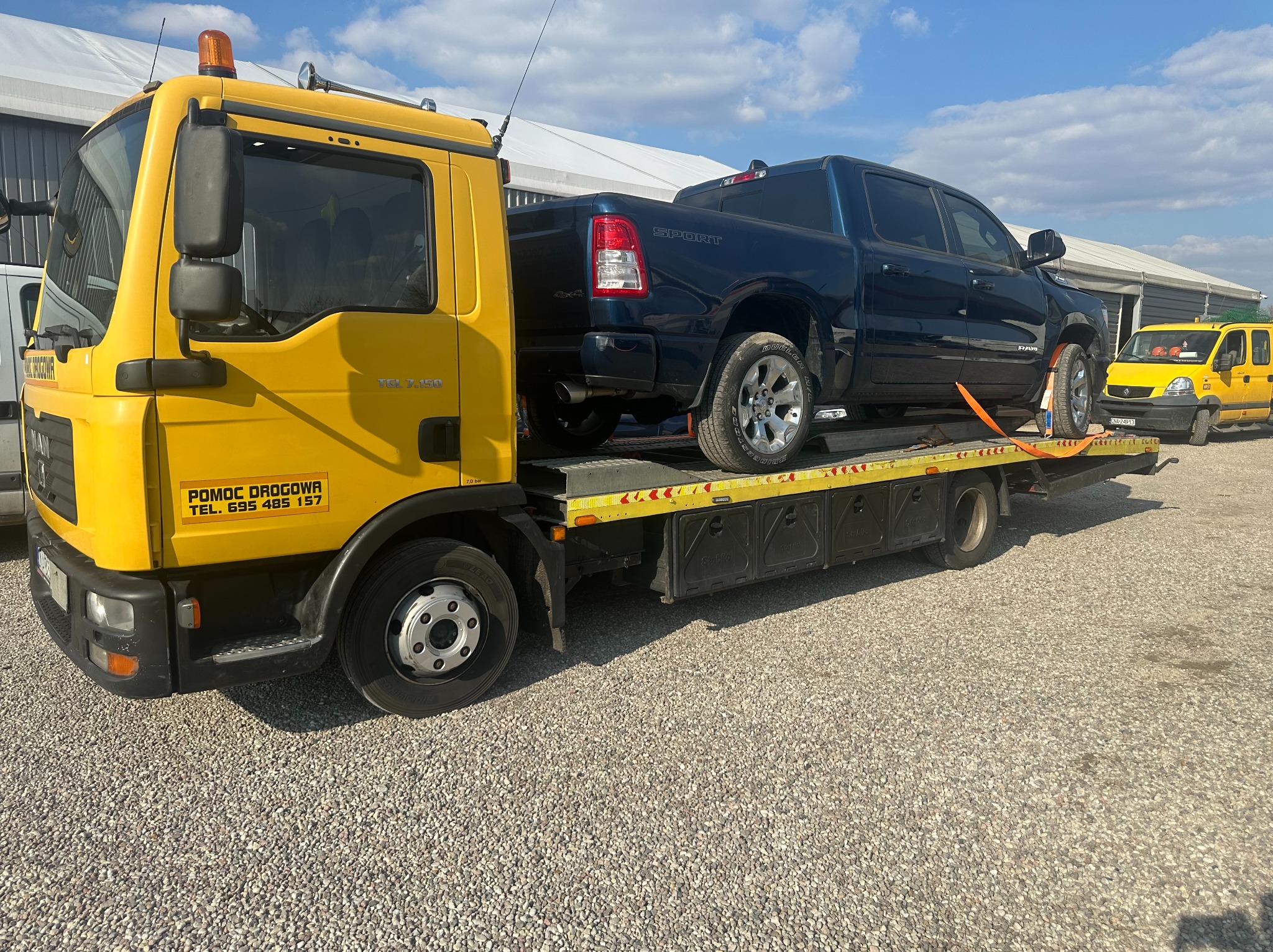 Żółta laweta MAN z niebieskim pickupem RAM na platformie, zabezpieczonym pomarańczowymi pasami. W tle budynek i żółty samochód pomocy drogowej.