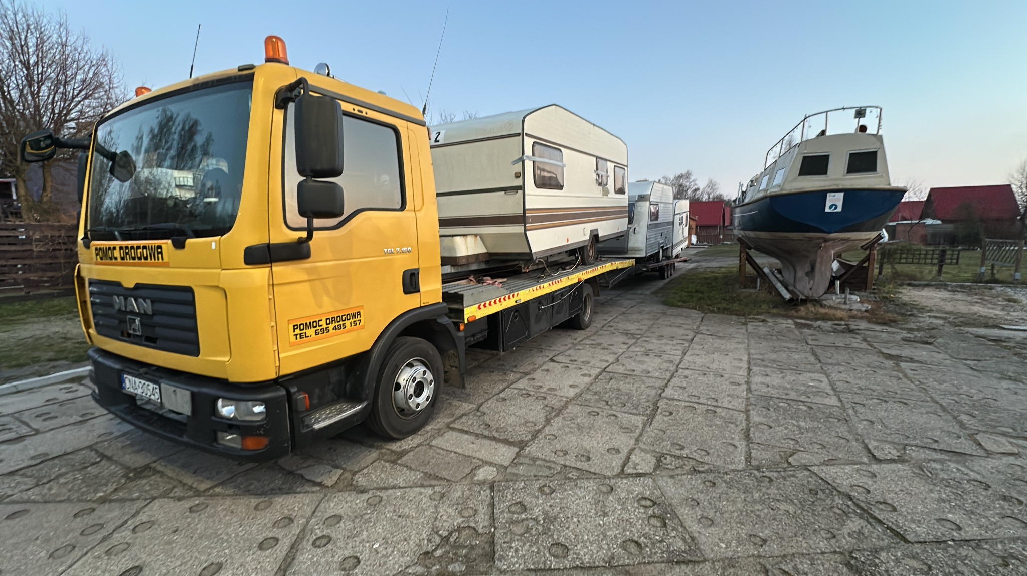 Żółty holownik MAN z przyczepą transportuje dwa białe kampery i łódź na betonowym placu. Widoczne budynki w tle.