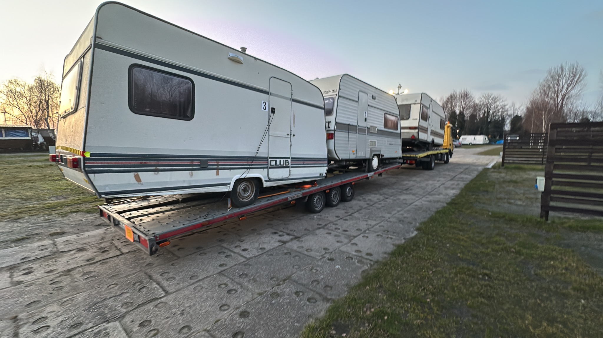 Laweta z trzema przyczepami kempingowymi w Nakle nad Notecią. Transport przyczep na niskopodwoziowej platformie. Widok z boku, w tle drzewa i ogrodzenie.