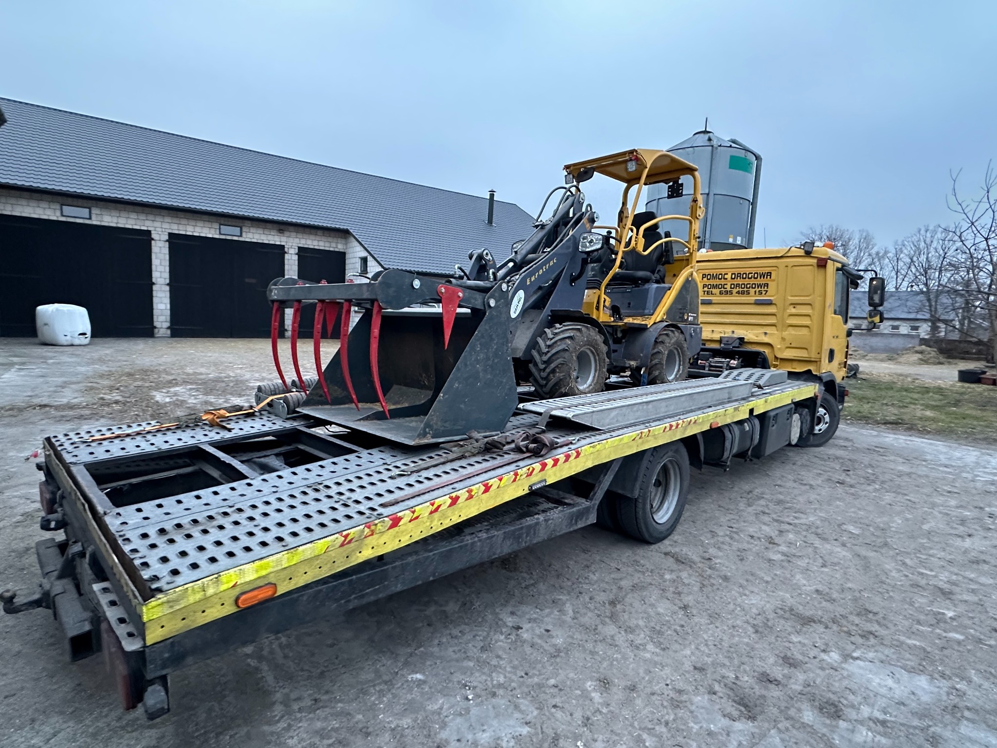 Żółta laweta z koparko-ładowarką Eurotrac na platformie. Widoczny budynek gospodarczy w tle. Pomoc drogowa z numerem telefonu na kabinie.