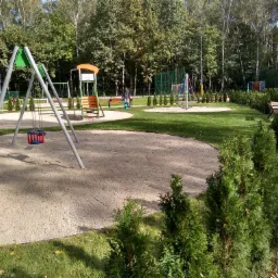 Osiedlowy plac zabaw z huśtawkami, zjeżdżalnią z koszem do koszykówki, karuzelą, ławkami i ścieżką z kostki brukowej, otoczony zielenią i drzewami.