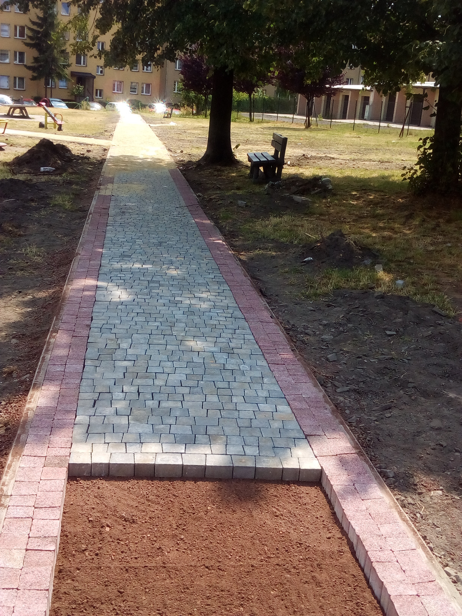 Fragment chodnika z kostki brukowej w trakcie układania, widoczny przygotowany obszar z podsypką, jasnoszare kostki w środku i czerwone obrzeża, w tle budynek mieszkalny i drzewa.