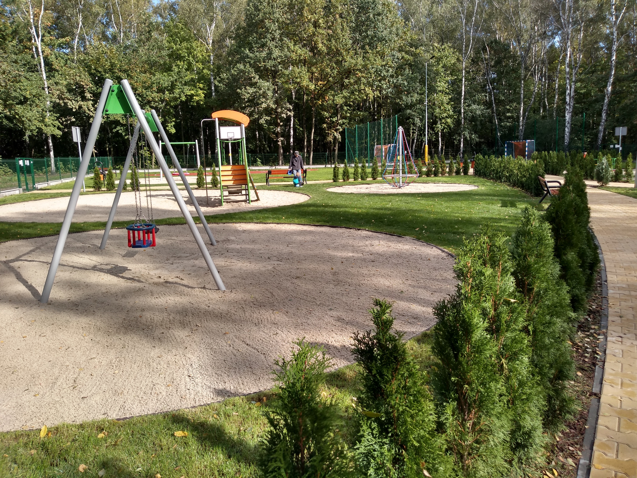 Osiedlowy plac zabaw z huśtawkami, zjeżdżalnią z koszem do koszykówki, karuzelą, ławkami i ścieżką z kostki brukowej, otoczony zielenią i drzewami.