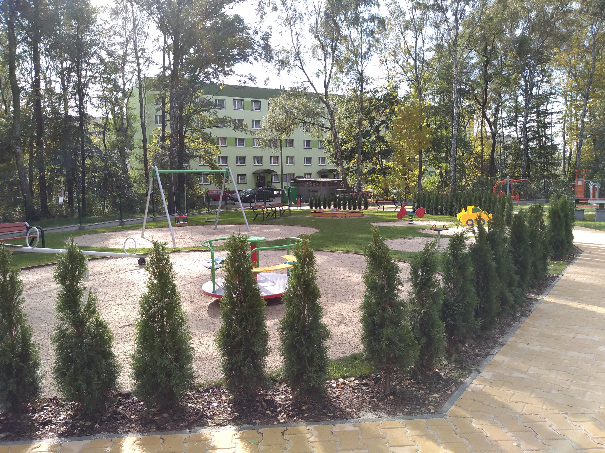 Osiedlowy plac zabaw w Halembie, Ruda Śląska, z huśtawkami, karuzelą i innymi urządzeniami, oddzielony od chodnika rzędem małych drzew iglastych, na tle zielonego bloku.