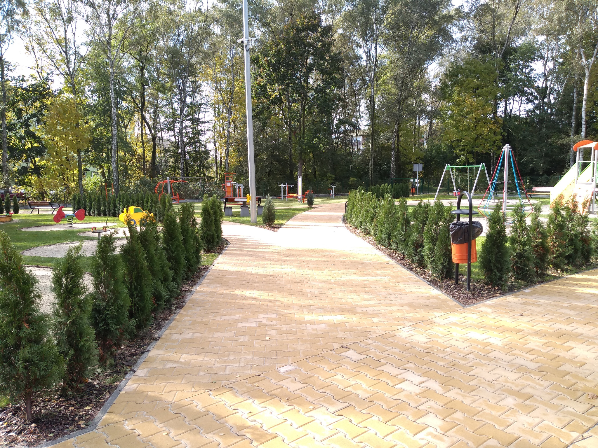 Żółta kostka brukowa ułożona w zygzak na ścieżce w parku, obrzeża wyznaczone przez niskie tuje, w tle plac zabaw z huśtawkami i innymi atrakcjami dla dzieci w Rudzie Śląskiej Halembie.