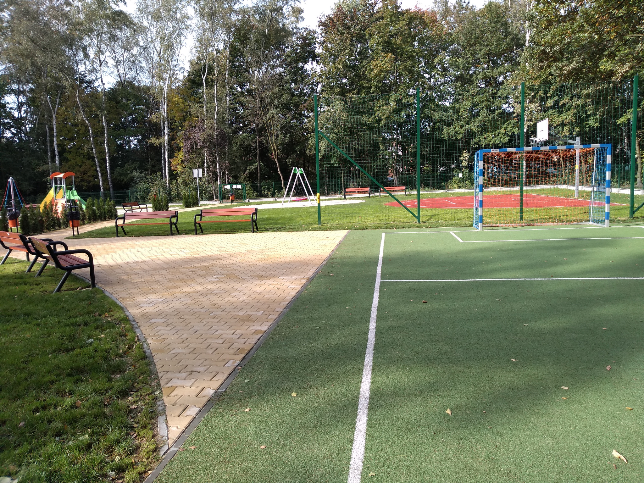 Plac zabaw w osiedlu Halemba z kostki brukowej prowadzącej do boiska z bramką, w tle huśtawki i zieleń parkowa.