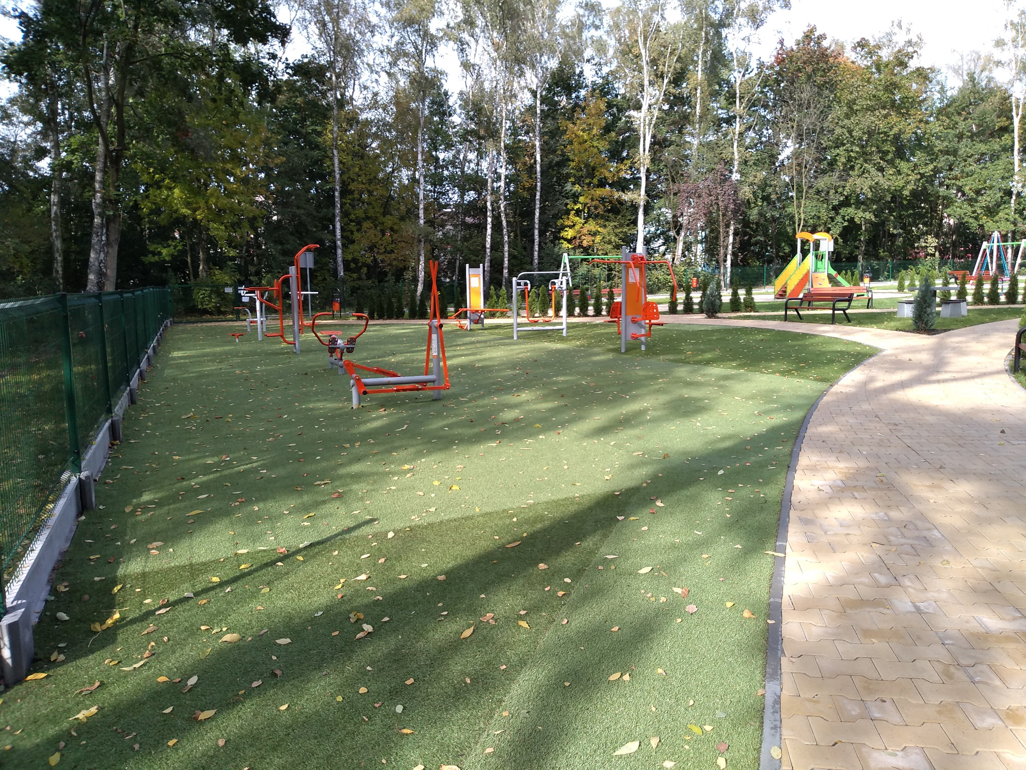 Osiedlowy plac zabaw w Rudzie Śląskiej - Halembie z zieloną, sztuczną nawierzchnią i zestawem pomarańczowo-szarych urządzeń do ćwiczeń na świeżym powietrzu, otoczony drzewami i ogrodzeniem z siatki.