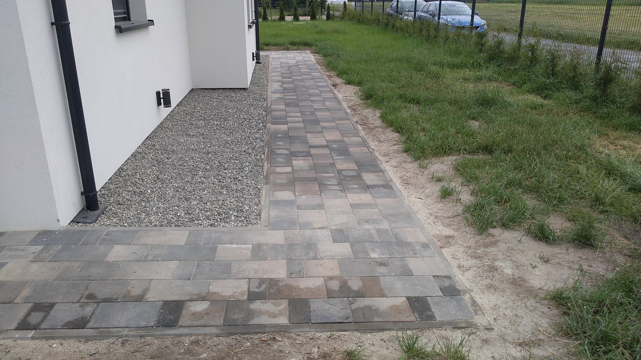 Ułożona ścieżka z kostki brukowej w odcieniach szarości prowadząca wzdłuż białego budynku, z fragmentem wysypanym drobnym żwirem i trawnikiem po prawej stronie.