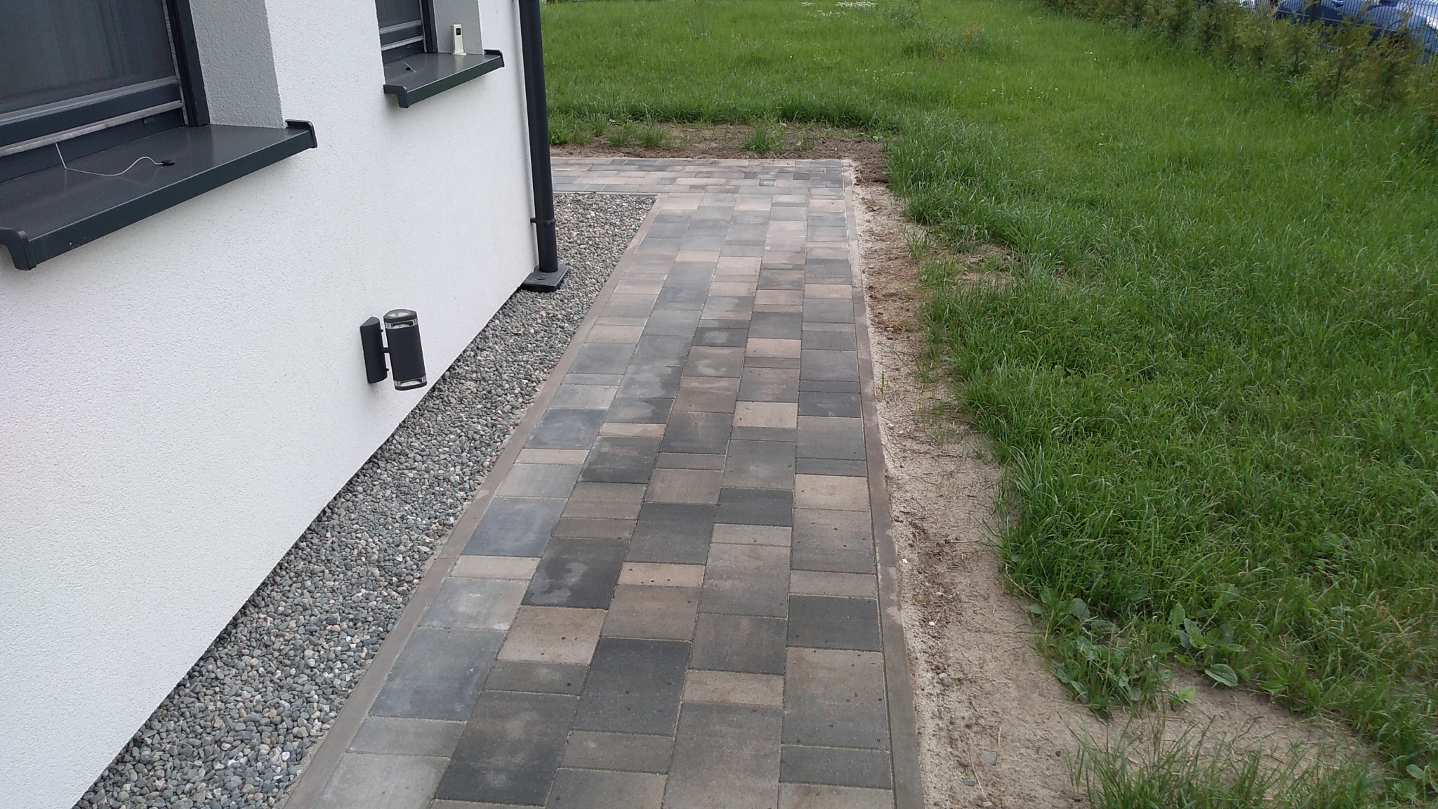 Ułożona ścieżka z kostki brukowej prowadząca wzdłuż ściany domu z widocznym oświetleniem zewnętrznym i pasem żwiru.