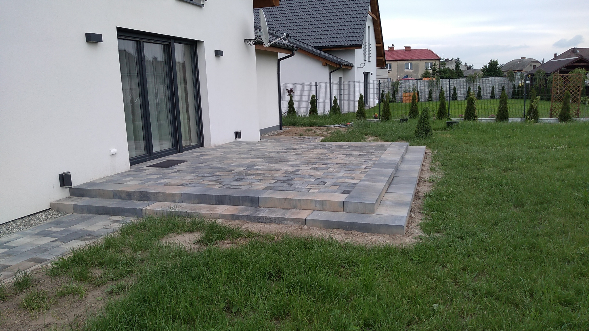 Ułożona kostka brukowa tworząca taras z dwoma stopniami przy białym domu z czarną stolarką okienną, widok na ogród z tujami wzdłuż metalowego ogrodzenia.