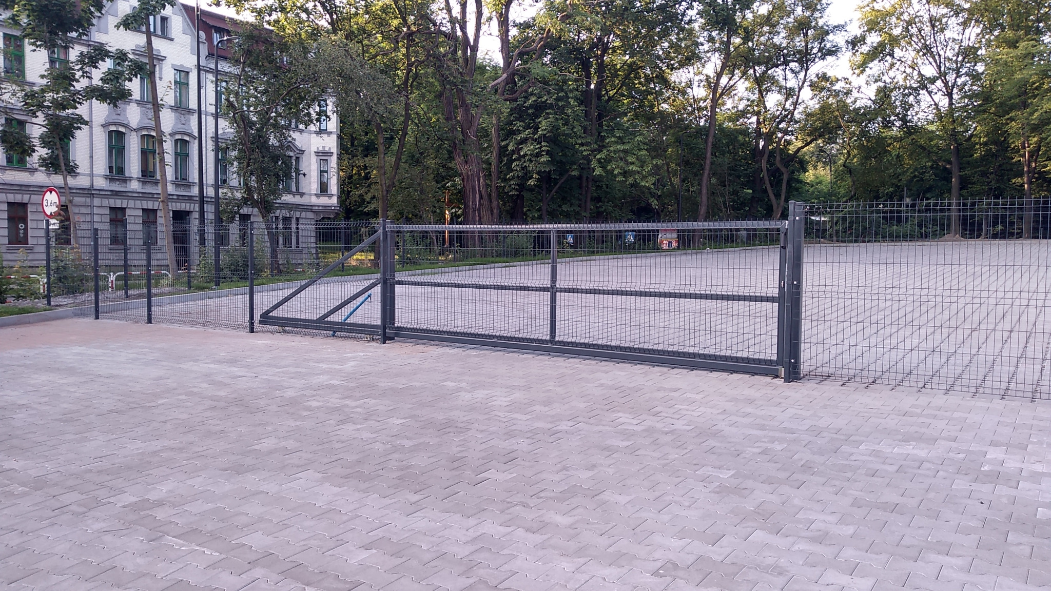 Przesuwna brama z siatki na parkingu z kostki brukowej w Rudzie Śląskiej, widoczny znak drogowy ograniczenia wysokości 3,6m oraz budynek w tle.