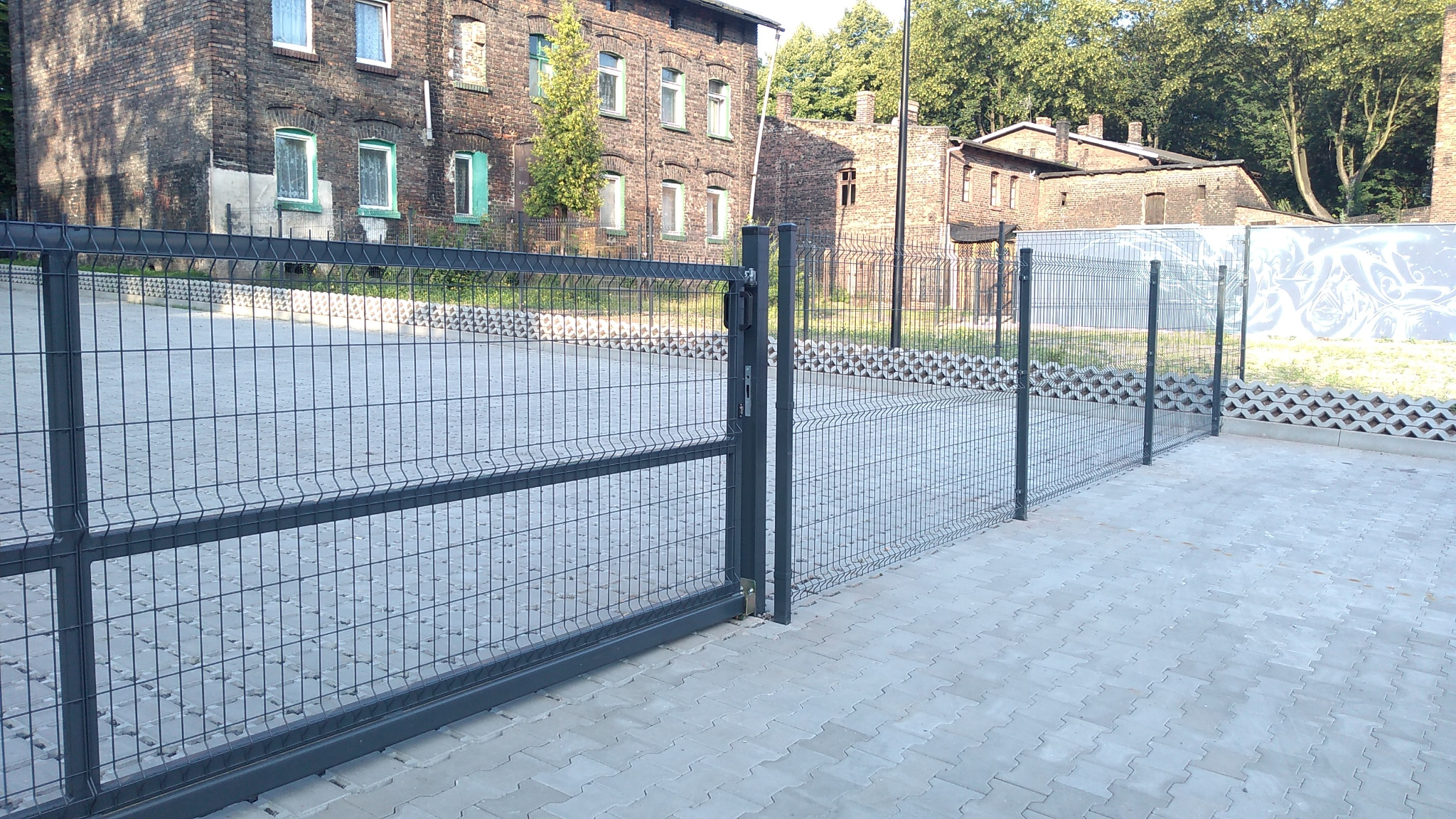 Nowoczesne, grafitowe ogrodzenie panelowe z bramą przesuwną, zabezpieczające parking z kostki brukowej, w tle ceglany budynek z zielonymi obramowaniami okien w Rudzie Śląskiej.