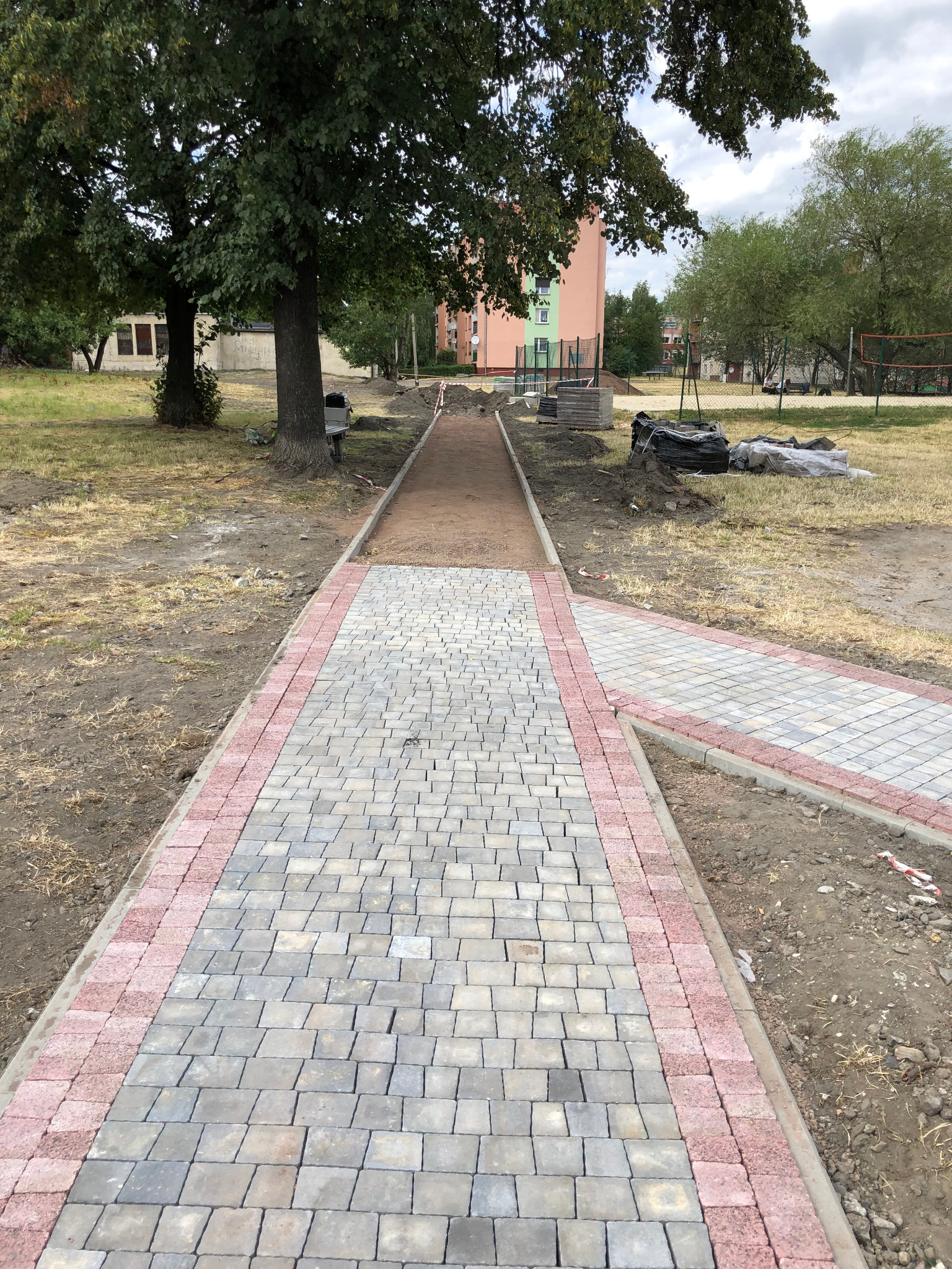 Nowo ułożony chodnik z szarej kostki brukowej z obrzeżem z czerwonej kostki, prowadzący przez park w Rudzie Śląskiej, widoczne przygotowane podłoże i elementy do dalszych prac.
