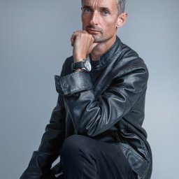 Studio Fotograficzne Laura Bąk - Mężczyzna w skórzanej kurtce, z zegarkiem na nadgarstku, siedzi na stołku opierając brodę na dłoni, patrzy w obiektyw na szarym tle.