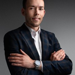 STUDIO FOTOGRAFII ARTYSTYCZNEJ. LAURA BĄK - Portret młodego mężczyzny w granatowej marynarce w kratę, białej koszuli i zegarku na metalowej bransolecie, stojącego na tle szarego tła z rękami skrzyżowanymi na piersiach.