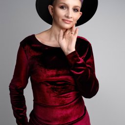 STUDIO FOTOGRAFII ARTYSTYCZNEJ. LAURA BĄK - Portret kobiety w bordowej welurowej sukience i czarnym kapeluszu, z krótkimi fioletowymi włosami, dłonią dotykającej policzka, pozującej na szarym tle.