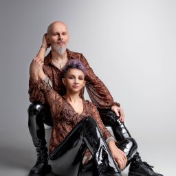 STUDIO FOTOGRAFII ARTYSTYCZNEJ. LAURA BĄK - Portret pary: mężczyzna z siwą brodą opiera rękę na ramieniu kobiety z krótkimi fioletowymi włosami, oboje w brązowych wzorzystych koszulach i czarnych lakierowanych spodniach, siedzą na białym tle.