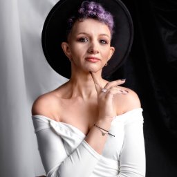 STUDIO FOTOGRAFII ARTYSTYCZNEJ. LAURA BĄK - Portret kobiety z fioletowymi włosami w czarnym kapeluszu, ubrana w białą sukienkę z odkrytymi ramionami, pozująca na tle białej i czarnej tkaniny.
