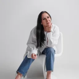 Portret kobiety siedzącej na białym krześle w obszernym białym męskim koszuli i dżinsach, z ręką podpartą pod brodą, na jasnoszarym tle.