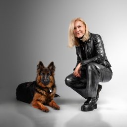 Studio Fotograficzne Laura Bąk - Kobieta w skórzanym stroju kuca obok owczarka niemieckiego na jasnoszarym tle. Profesjonalne oświetlenie podkreśla detale stroju i sierści psa. Uśmiech kobiety dodaje ciepła.