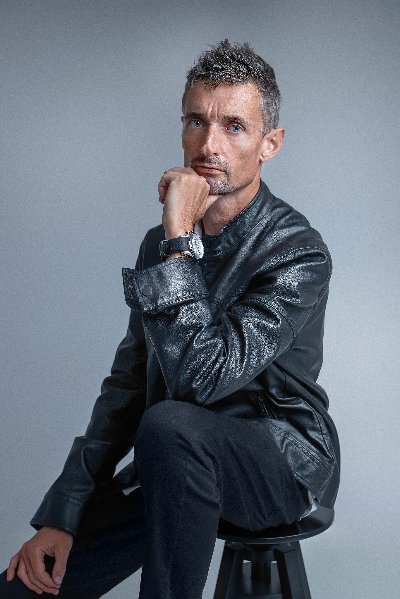 Mężczyzna w skórzanej kurtce, z zegarkiem na nadgarstku, siedzi na stołku opierając brodę na dłoni, patrzy w obiektyw na szarym tle.