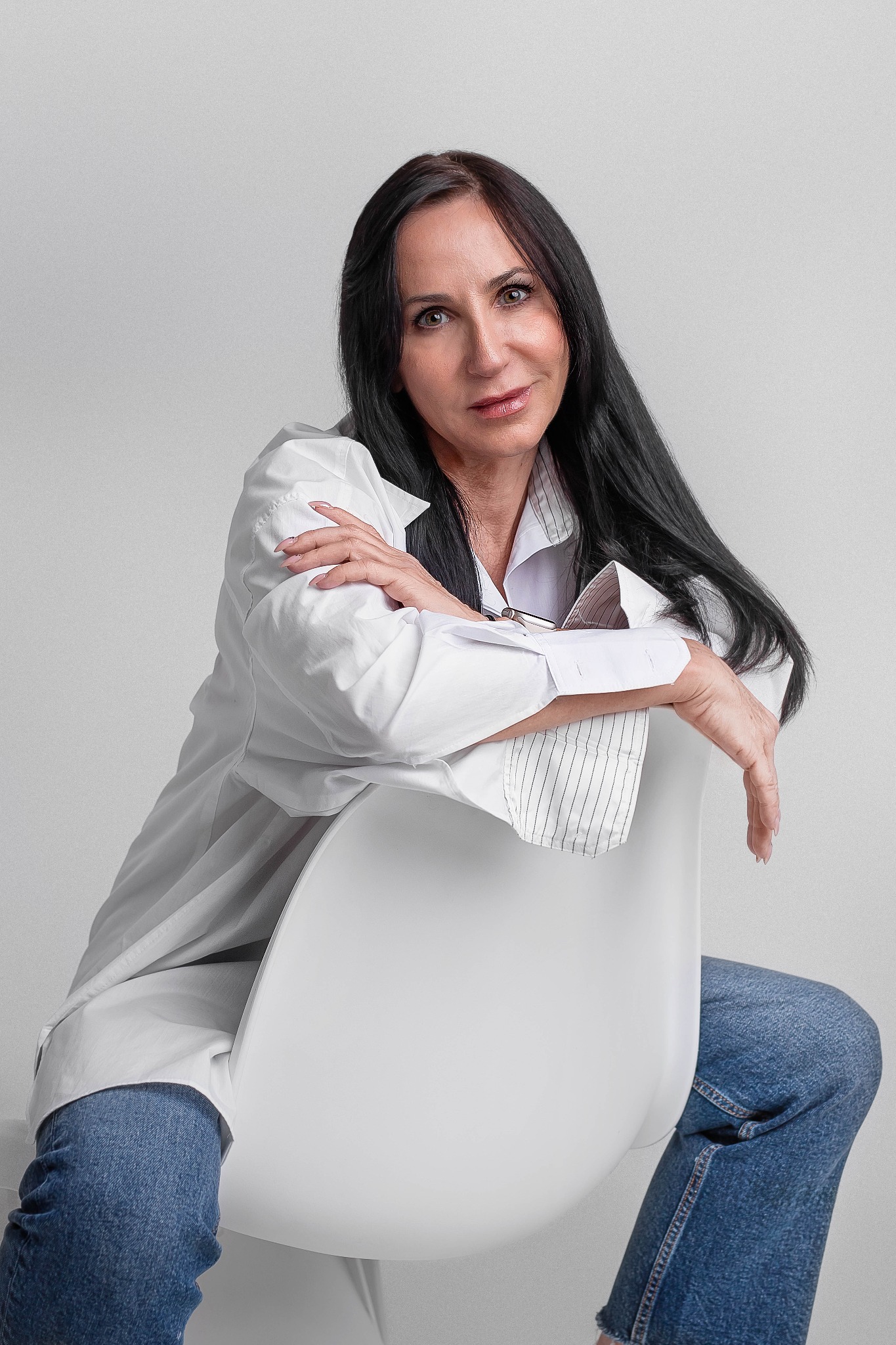 Portret kobiety z długimi, czarnymi włosami, w białej koszuli i dżinsach, siedzącej na białym krześle na neutralnym tle.