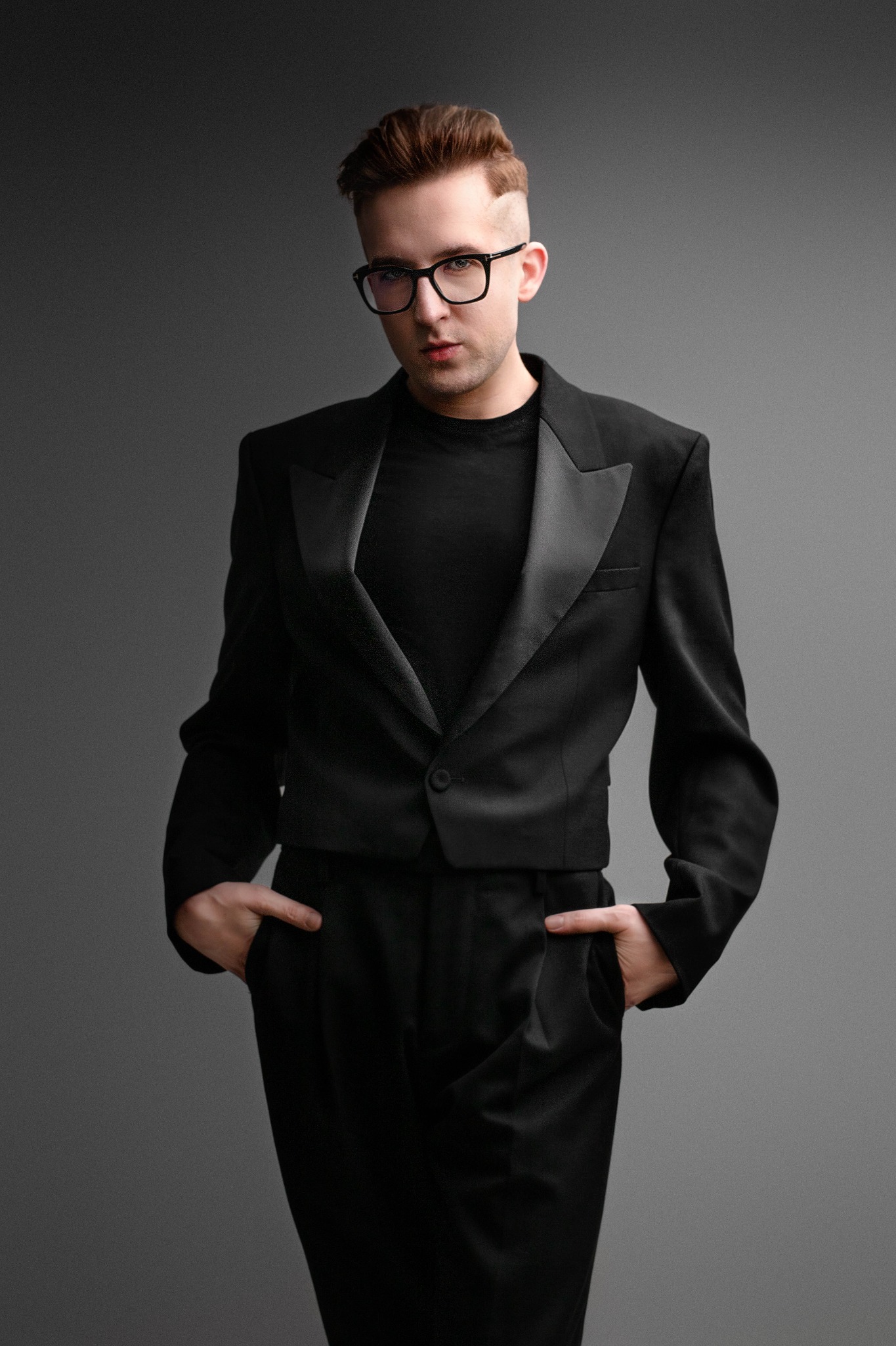 Mężczyzna w czarnym, krótkim smokingu i okularach, pozujący na szarym tle z rękami w kieszeniach.